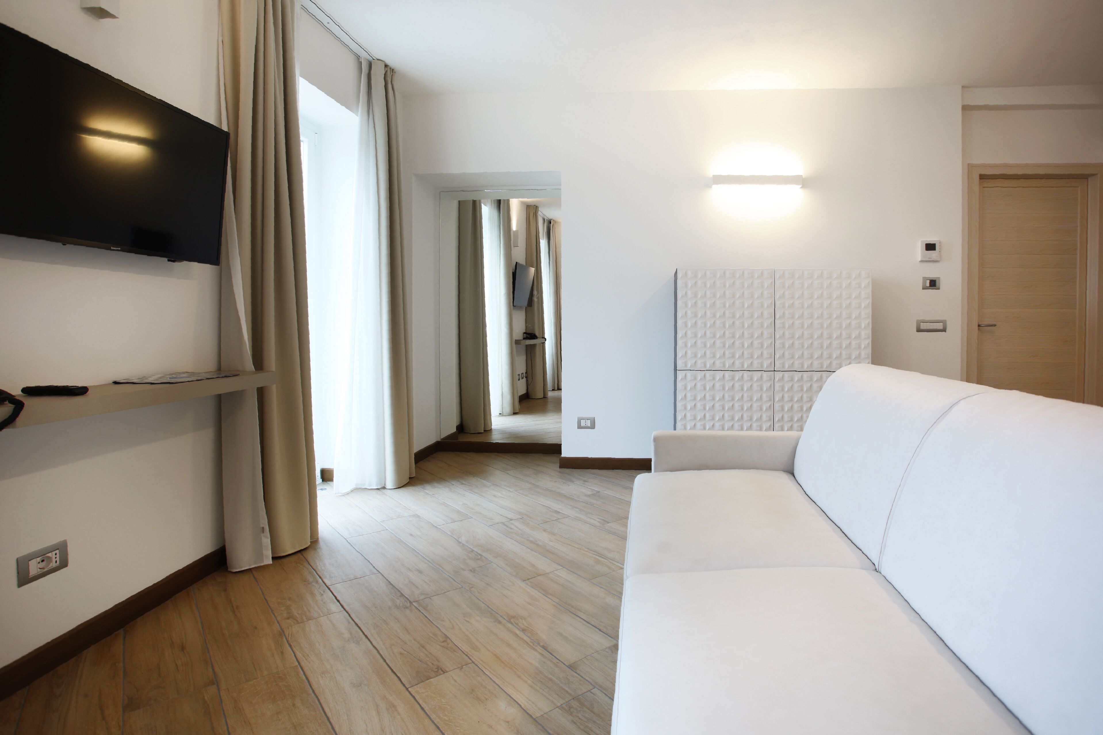 Een moderne hotelkamer met een witte bank, een flatscreen televisie aan de muur, een houten vloer en uitzicht op de gang.
