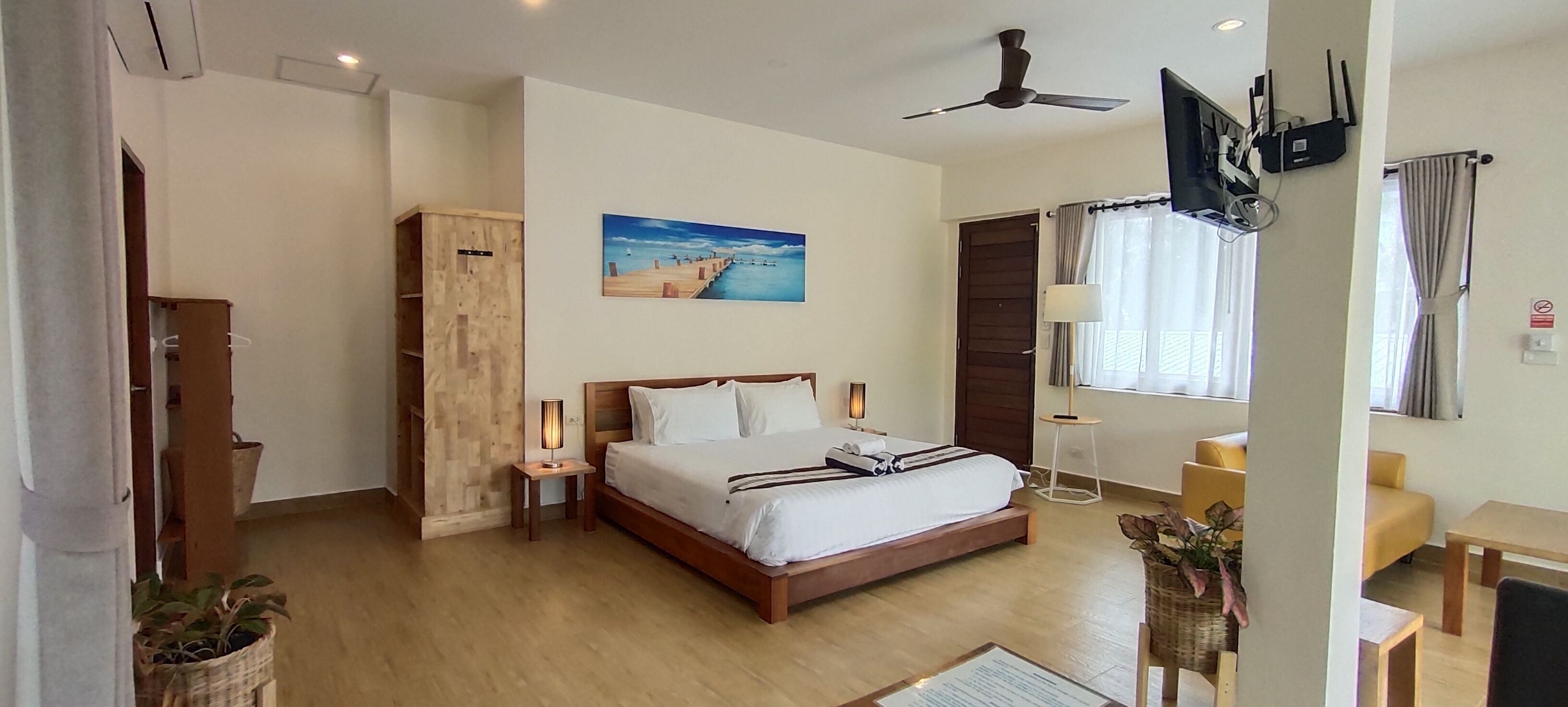 A hotel room with a wooden bed, a nightstand with a lamp, a desk, a chair, a window with curtains, a painting on the wall, and a ceiling fan.