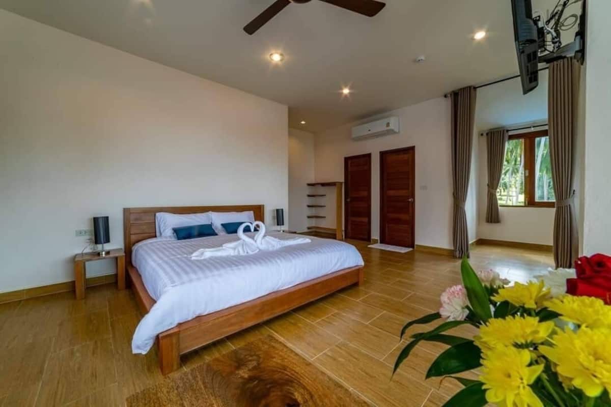 Un dormitorio amplio con una cama grande, piso de madera, ventilador de techo y vista al exterior a través de una ventana.