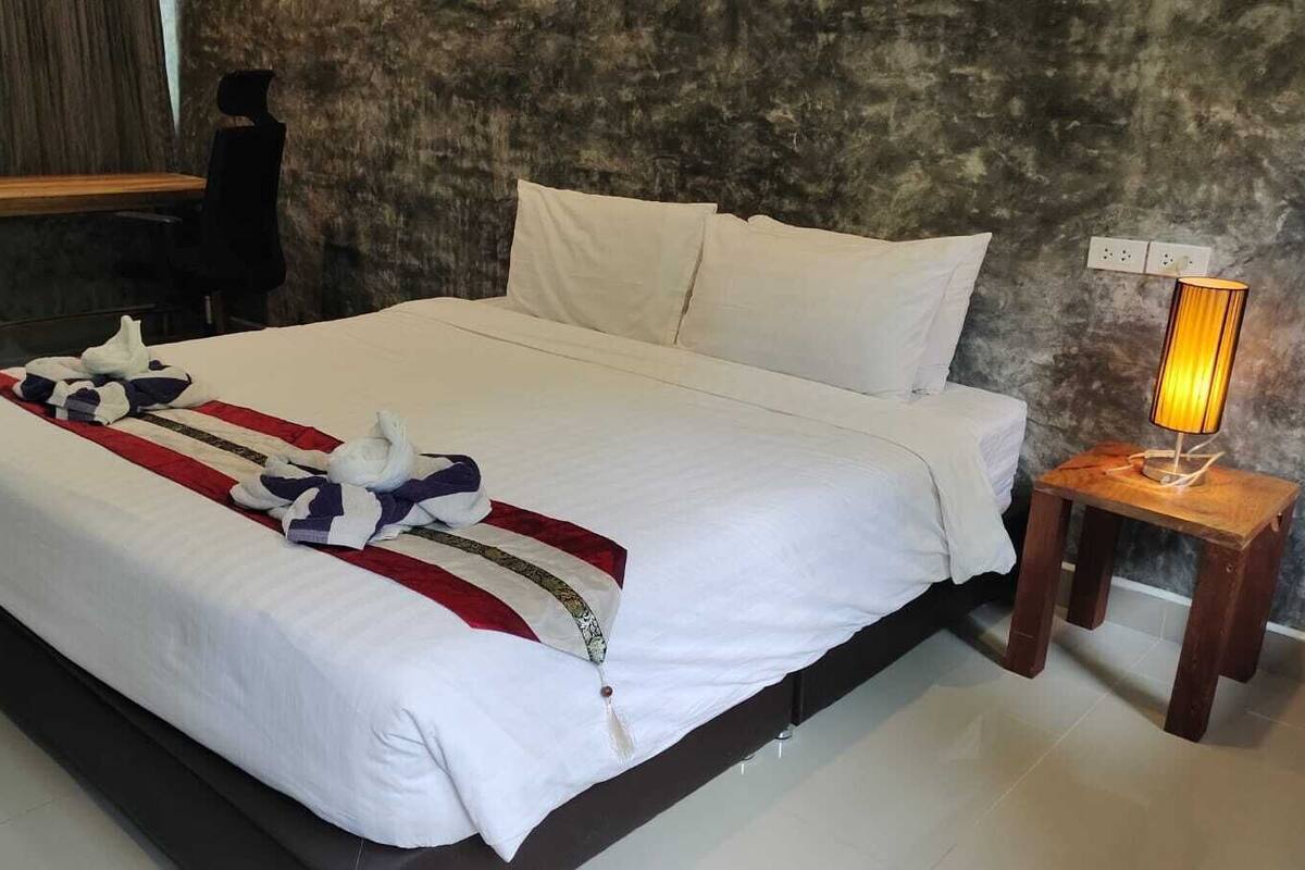 Una cama bien tendida con sábanas blancas y una manta a rayas, una mesita de noche con lámpara y un cabecero de madera oscura.