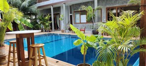 A pool area with a wooden table and stools, surrounded by potted plants and a building with large windows.