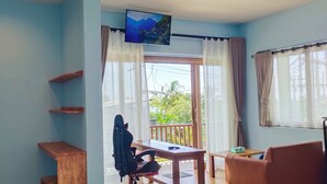 Una habitación con televisor montado en la pared, un escritorio de madera, una silla y un sofá. Hay dos ventanas con cortinas, y un balcón con barandilla.