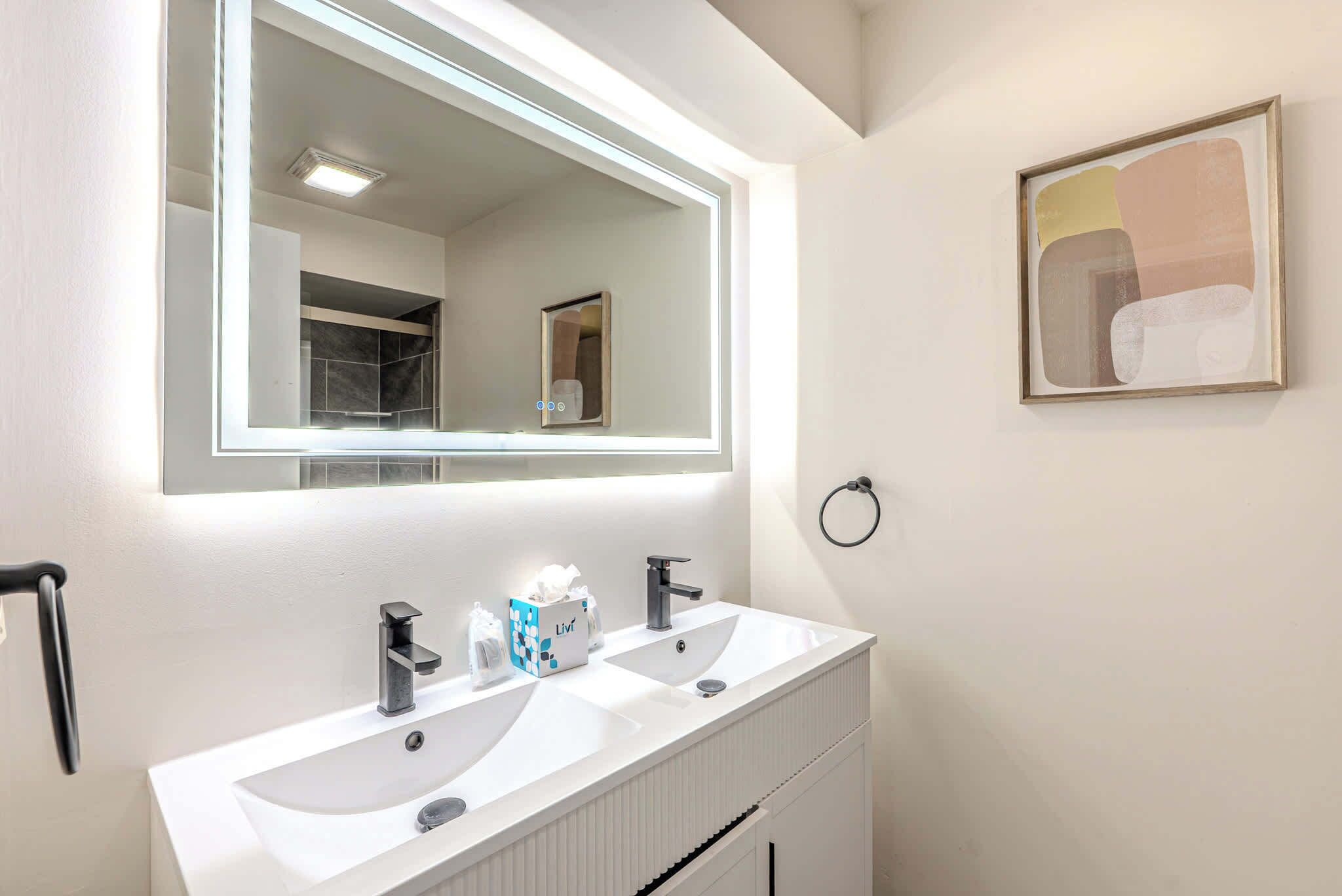 Une salle de bain avec deux lavabos, un grand miroir et un tableau abstrait encadré accroché au mur.