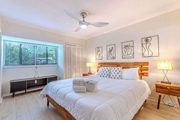 A bedroom with a bed, ceiling fan, artwork on the wall, bedside lamps, and a window with blinds.