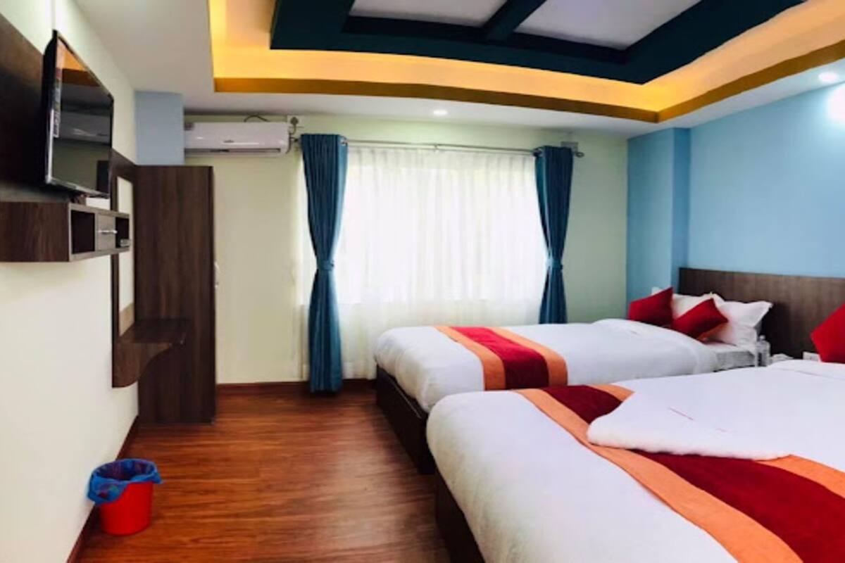 Ein Hotelzimmer mit zwei Betten, einem an der Wand befestigten Fernseher, Holzboden und einem Fenster mit Vorhängen.