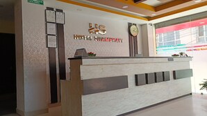 Un comptoir d’accueil d’hôtel avec une horloge, des certificats encadrés et une enseigne indiquant « HÔTEL SHAMBEHAVI ».