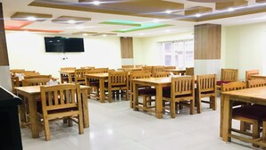 Un espace repas comprenant plusieurs tables et chaises en bois, un téléviseur à écran plat fixé au mur et des fenêtres équipées de stores.