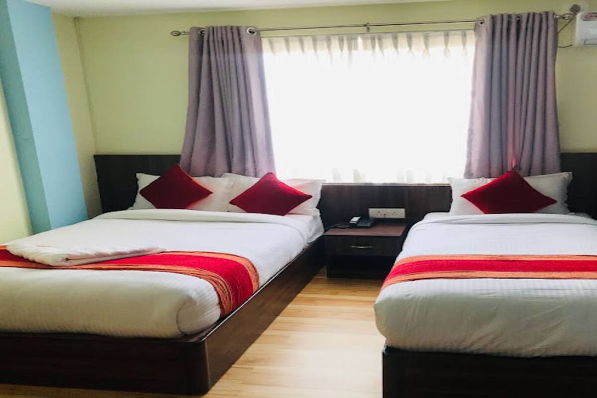 Ein Hotelzimmer mit zwei Einzelbetten, jedes mit einem roten Kissen und einer weißen Decke. Dazwischen befindet sich ein Nachttisch mit einer Lampe und einem Telefon. Das Zimmer verfügt über ein Fenster mit violetten Vorhängen und eine Klimaanlage darüber.