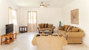 Ein Wohnzimmer mit Fernseher auf einem Holzboden, zwei Sofas mit gemusterten Kissen, einem Couchtisch und einem Deckenventilator.