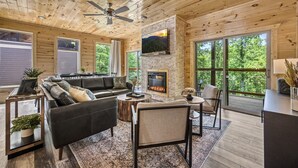 A spacious living room with a fireplace, large windows, and a ceiling fan. - Luxe Cabin|Indoor Pool~Game & Theater Rooms! (Sevierville)