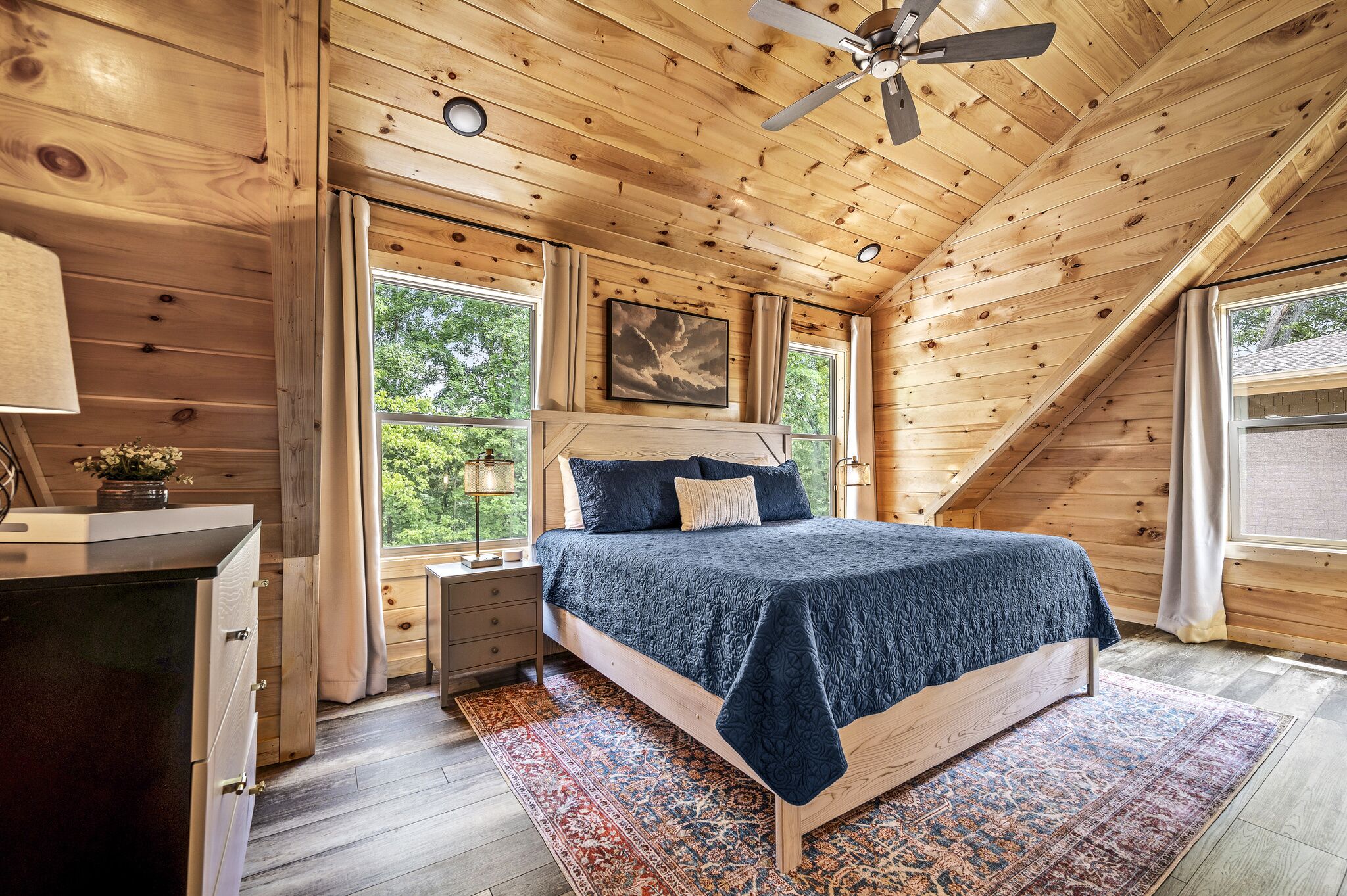 A bedroom with a wooden ceiling, a bed with a dark blue bedspread, a nightstand, a dresser, and a window with curtains.