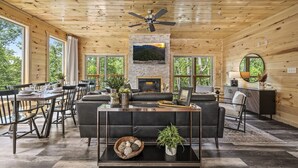 A spacious living room with a fireplace, a ceiling fan, and large windows offering a view of the outdoors. - Luxe Cabin|Indoor Pool~Game & Theater Rooms! (Sevierville)