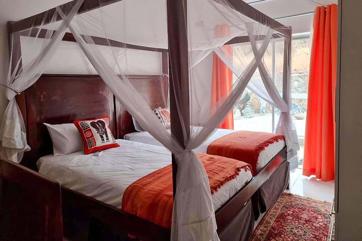 A four-poster bed with white and red bedding, a wooden headboard, and a view of the outdoors through a window with orange curtains.