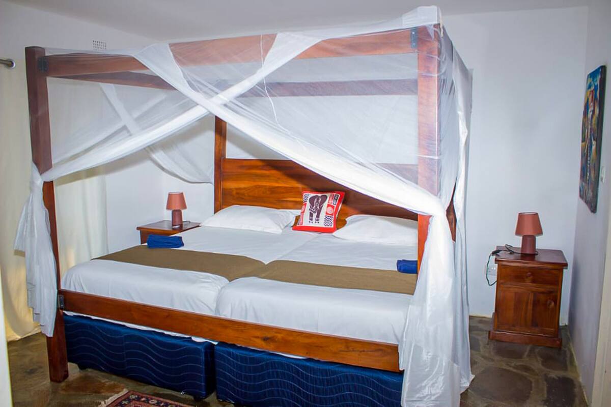 A wooden four-poster bed with a mosquito net, two bedside tables with lamps, and a small cabinet.