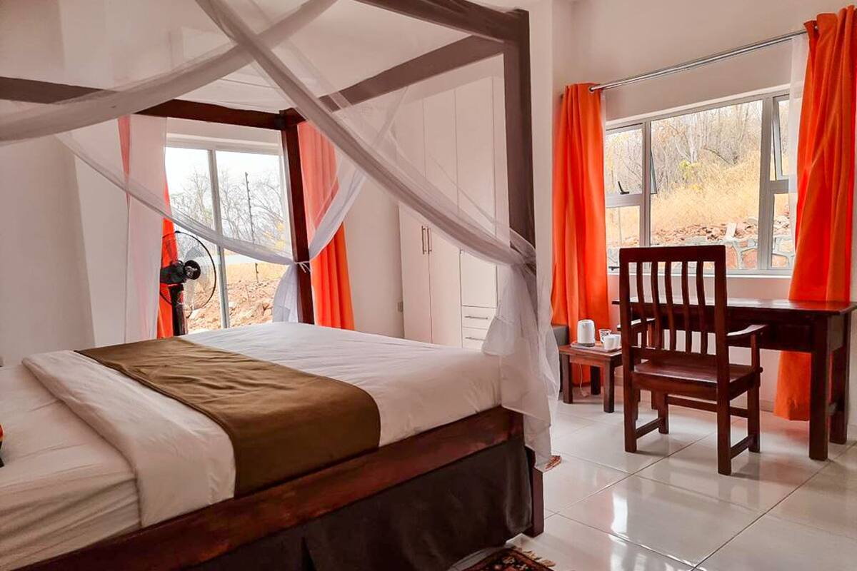 A bedroom with a four-poster bed, a desk with a chair, and a view of the outdoors through a window.