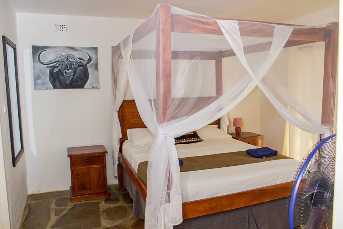 A room with a four-poster bed, a nightstand, a bedside table, a wall-mounted artwork, and a standing fan.