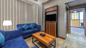 Moderni hotellihuone, jossa on sininen sohva, puinen sohvapöytä, litteänäyttöinen televisio ja näkymä ulkoiselle parvekkeelle.