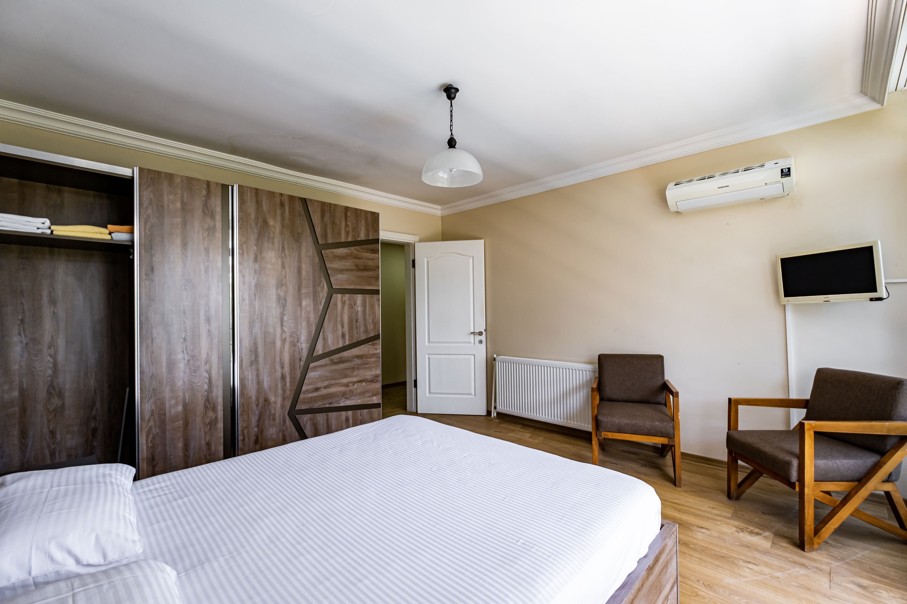 Quarto de hotel com uma cama, duas cadeiras de madeira, uma TV, ar condicionado e um guarda-roupa.