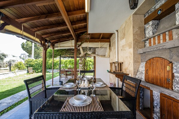 An outdoor dining area with a glass table, wicker chairs, and a stone fireplace.