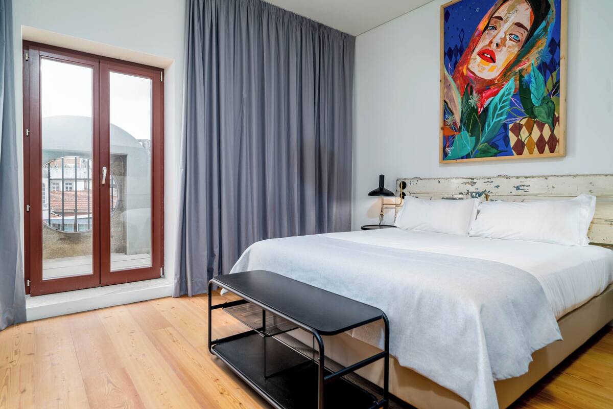 Un dormitorio con cama, una banca, un cuadro y vistas por una ventana.