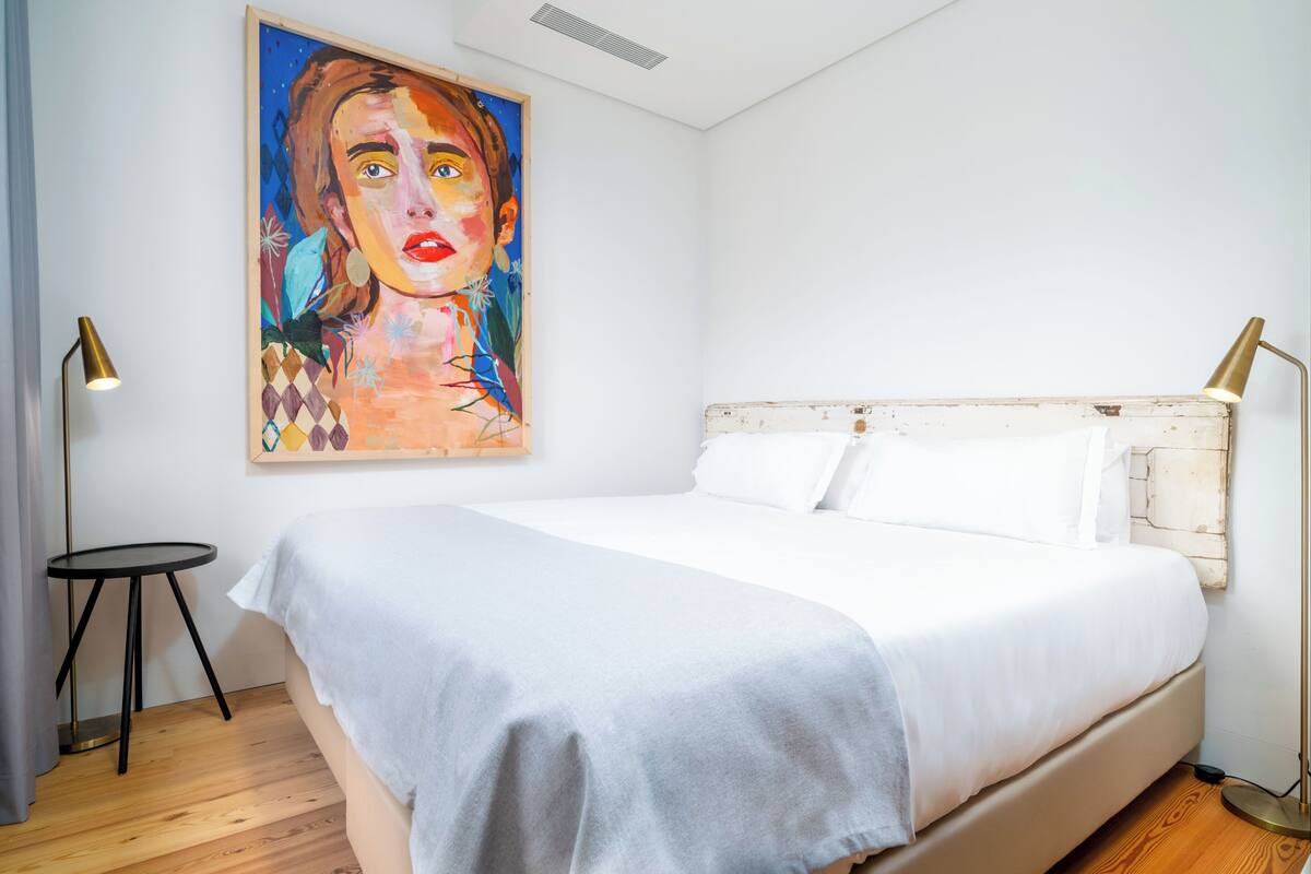 Un dormitorio con una cama grande, una mesita redonda pequeña, dos lámparas de pie y un cuadro colorido en la pared.
