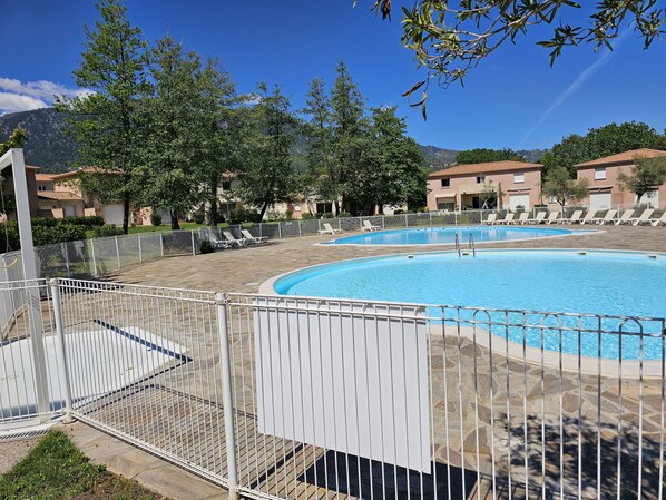 A swimming pool with a white fence, lounge chairs, and buildings in the background.