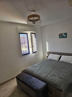 A bedroom with a bed, a bench, a window with curtains, a ceiling fan, and a wall-mounted light fixture.