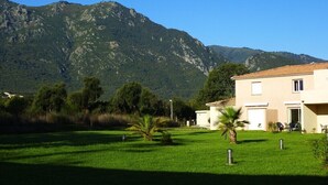 A building with a flat roof and a small attached structure, surrounded by a well-maintained lawn and palm trees, with mountains in the background. - Villa Melody Calme DS Residence Securisee Avec Piscines Proche MER ET Montagne (Santa-Maria-Poggio)