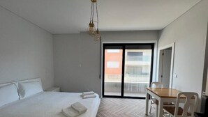 A modern hotel room with a large bed, a dining table, and a sliding glass door leading to a balcony.