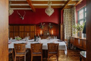 Ein Esszimmer mit roter Wand, Holzvertäfelung und einer Chandelier. Vier Holzstühle mit Leopardenmuster-Kissen, ein für vier Personen gedeckter Tisch und ein Vintage-Radio an der Wand.