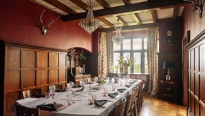 A dining room with a long table set for a meal, wooden paneling, chandeliers, and a window with curtains. - Christie Mansion - Luxury Cottages (Shanklin)