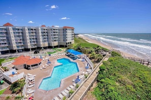 Un resort con una gran piscina, acceso a la playa y un edificio de varios pisos con balcones.