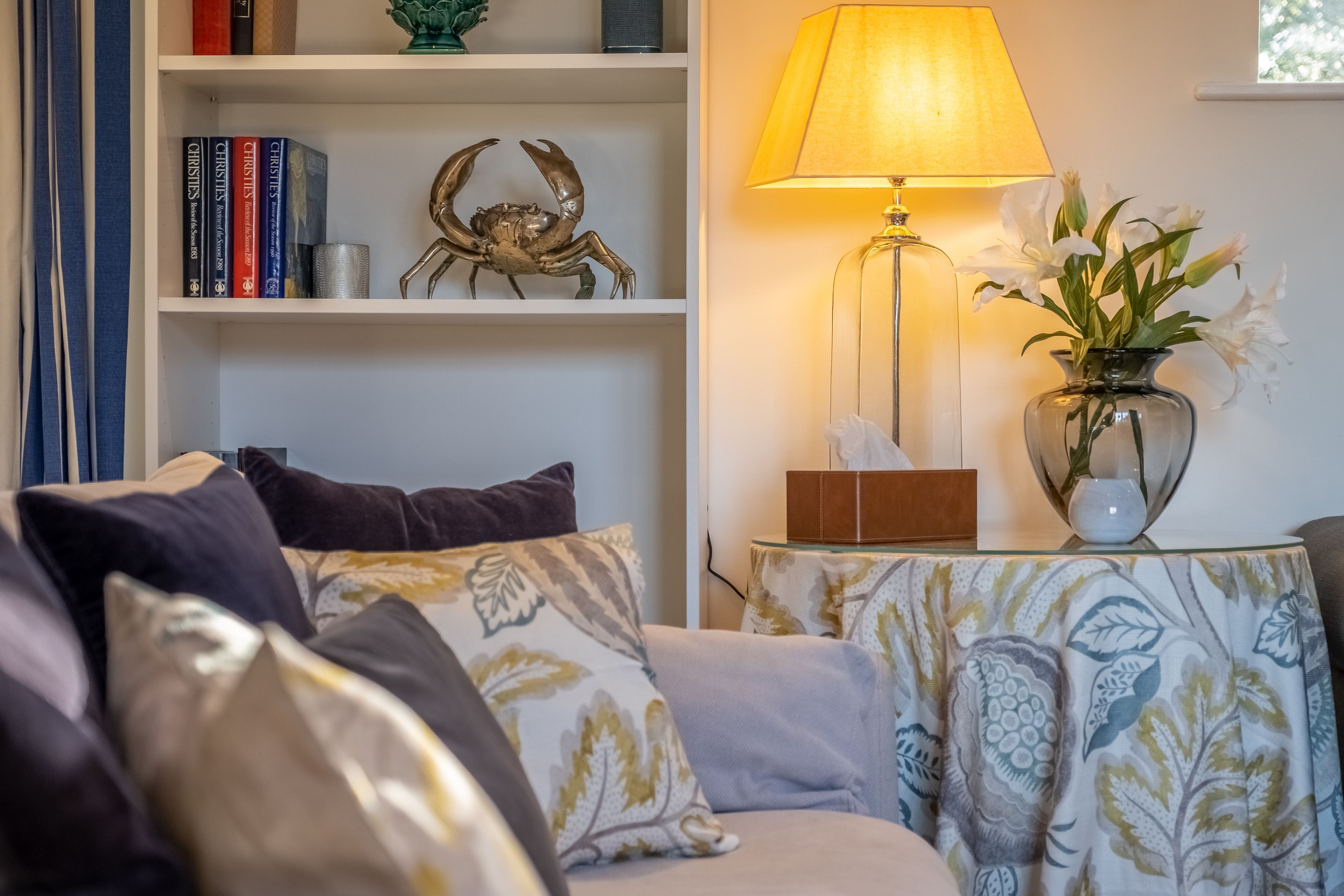 A cozy living room with a sofa, patterned cushions, a bookshelf with books and a decorative crab, a table with a lamp and vase of flowers.