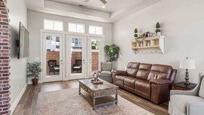 A spacious living room with a brown leather sofa, a wooden coffee table, and a white armchair. There is a ceiling fan, a TV mounted on the wall, and a small table with a lamp. - The Ella:  Charming 1-level private home, community amenities. (Hurricane)