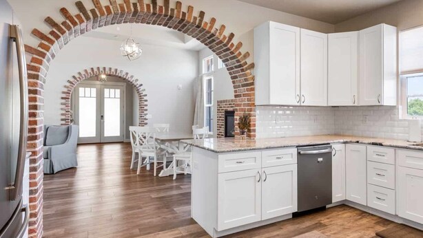 A modern kitchen with white cabinets, a brick archway, and a dining area with a table and chairs. - The Ella:  Charming 1-level private home, community amenities. (Hurricane)