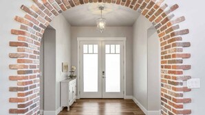 A hallway with a brick archway, white double doors, a wooden floor, a white console table with a vase, and a hanging light fixture. - The Ella:  Charming 1-level private home, community amenities. (Hurricane)