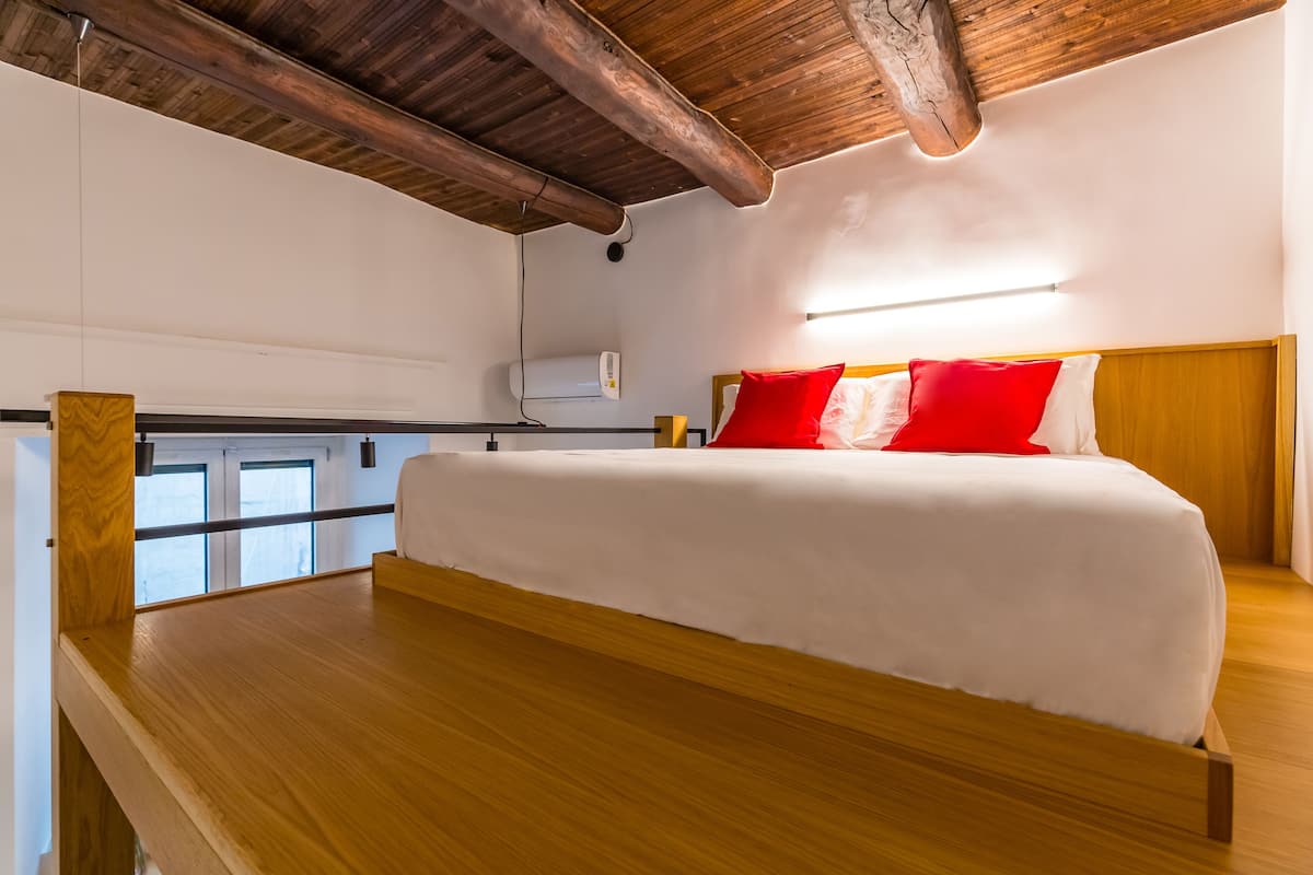 A bedroom with a wooden bed, white bedding, and red pillows. There is a ceiling fan, a window with blinds, and a wall-mounted air conditioner.