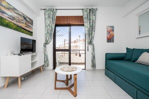 A modern living room with a white TV stand, a teal sofa, a round table, and a view through a sliding door. - 1 bedroom flat in Manta Rota by AlgarveManta (9T1) (Manta Rota)