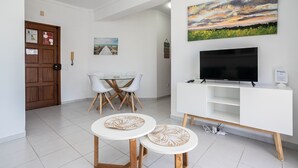 Una sala moderna con un mueble blanco para el televisor, un televisor de pantalla plana, una mesa de comedor con sillas y un cuadro en la pared.