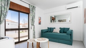 Una habitación con un sofá color turquesa, dos mesitas redondas, un ventanal con cortinas, un aire acondicionado blanco y un espejo en la pared.