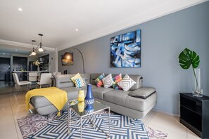 A modern living room with a grey sofa, colorful pillows, a blue abstract painting, and a large green plant.