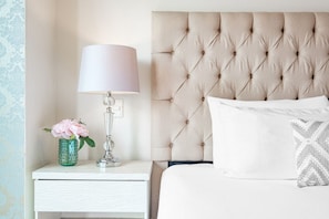 Un lit bien fait, avec une tête de lit capitonnée, une lampe de chevet, un vase décoratif avec des fleurs et un coussin à motifs.