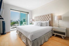 Habitación de hotel con una cama grande, una mesita de noche, una lámpara y vistas a la ciudad a través de un ventanal amplio.