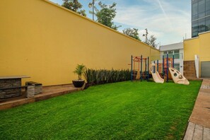 Un espace extérieur bien entretenu, avec un mur jaune, une pelouse verdoyante et des jeux pour enfants.