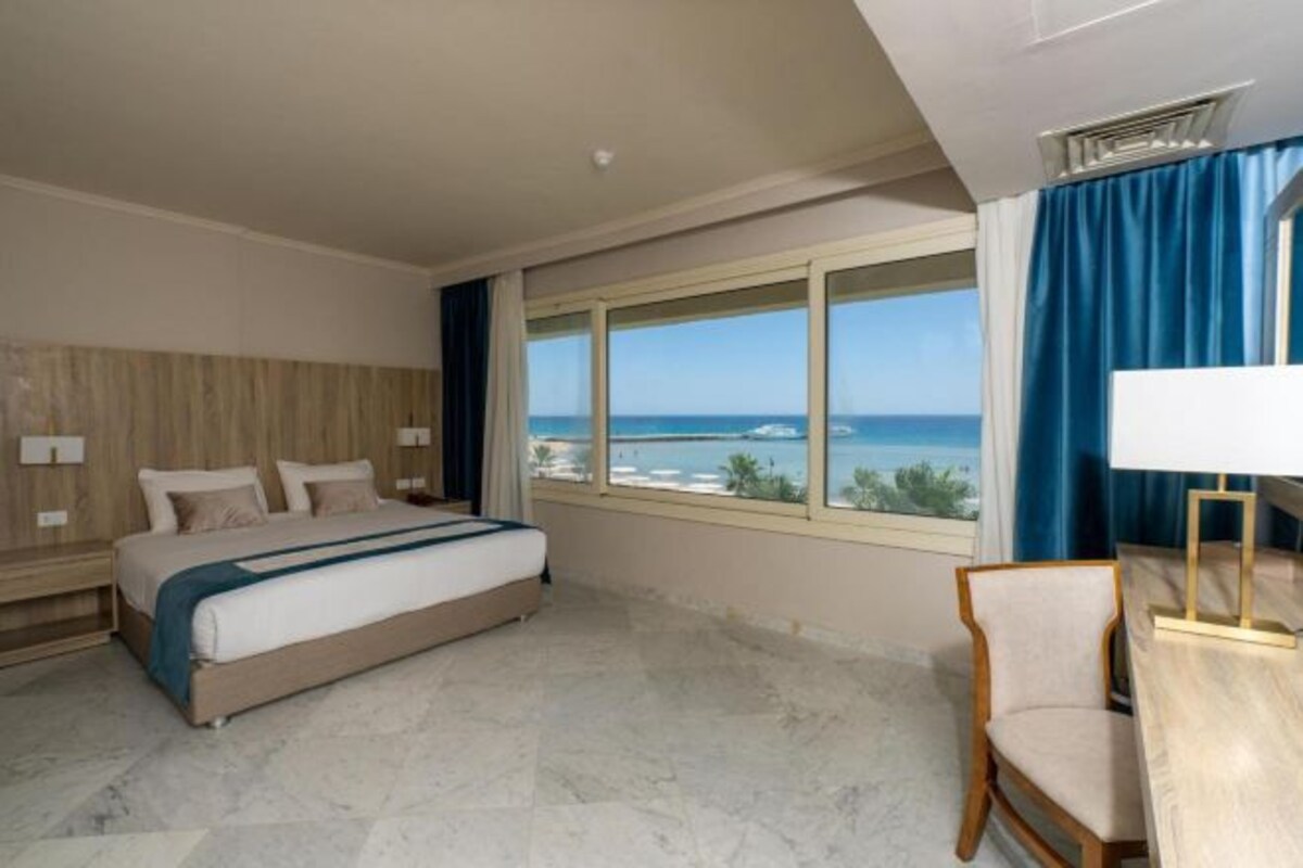 A hotel room with a bed, a desk, a chair, a window with a view of the sea, and a curtain.