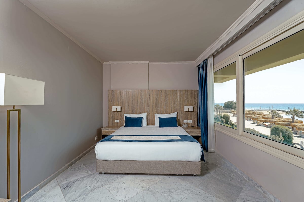 A hotel room with a large bed, a nightstand, a lamp, and a view of the beach through a large window.