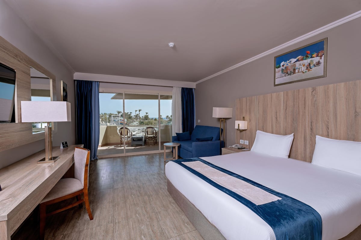 A hotel room with a large bed, a desk with a chair, a small table, and a view of the outdoors through a sliding door.