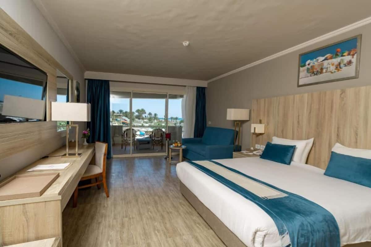 A hotel room with a large bed, a desk, a chair, and a balcony with a view of the city.