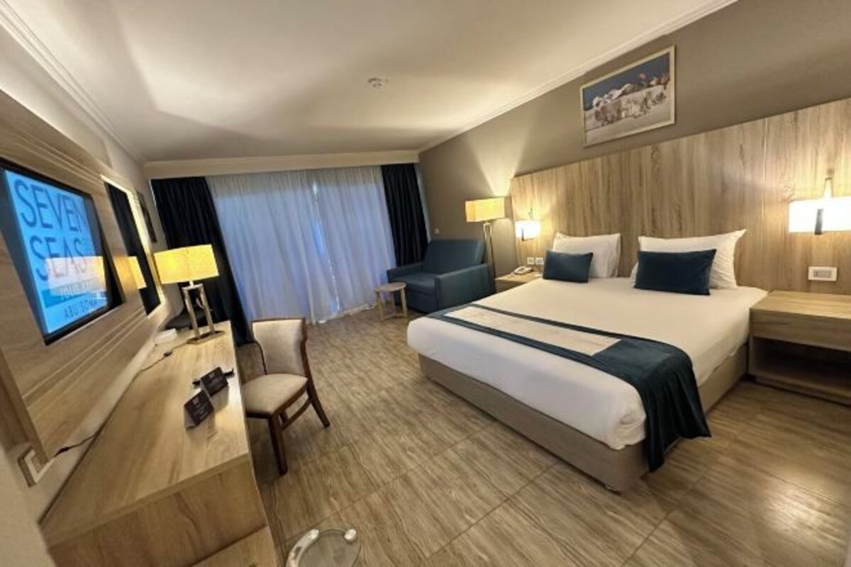 A hotel room with a large bed, a desk with a computer, a chair, and a TV displaying 'SEVEN SEAS'.