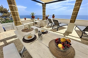 A terrace with a table set for two, featuring plates, cutlery, and a bowl of fruit. There are potted plants and a view of the sea.